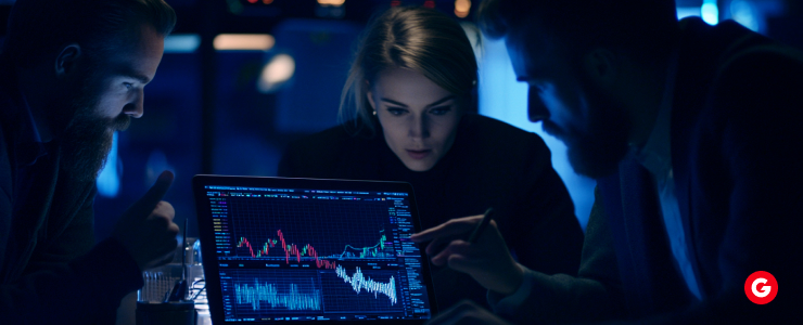 Three individuals observe a computer screen displaying a stock market graph, engaged in analysis and discussion.